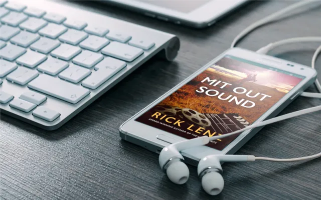 Audiobook version of Mit Out Sound is displayed on a smartphone with earbuds plugged in while sitting next to a keyboard.